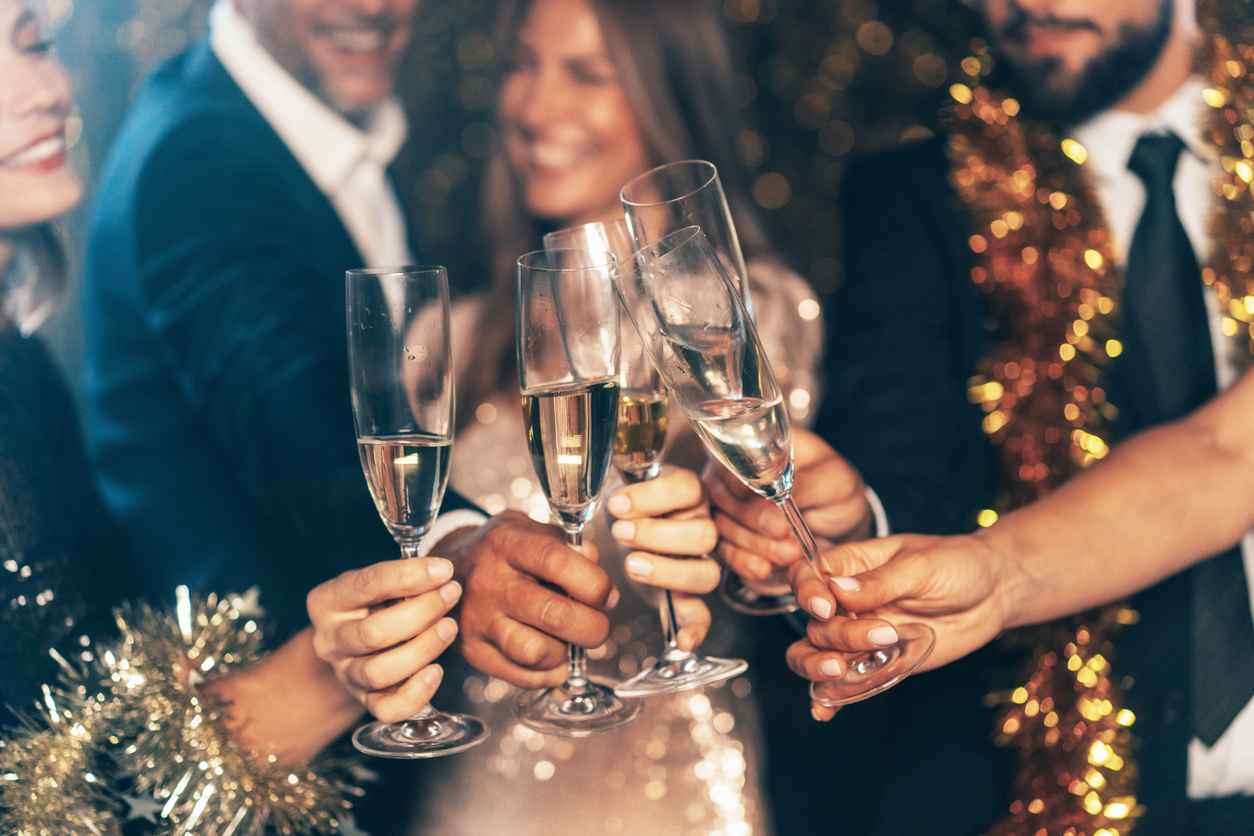 Friends clinking champagne glasses. Group of elegant friends celebrating New year or Christmas. Mid adult beautiful women in sparkle dress and stylish men dancing and holding glasses of champagne and smiling; corporate holiday party