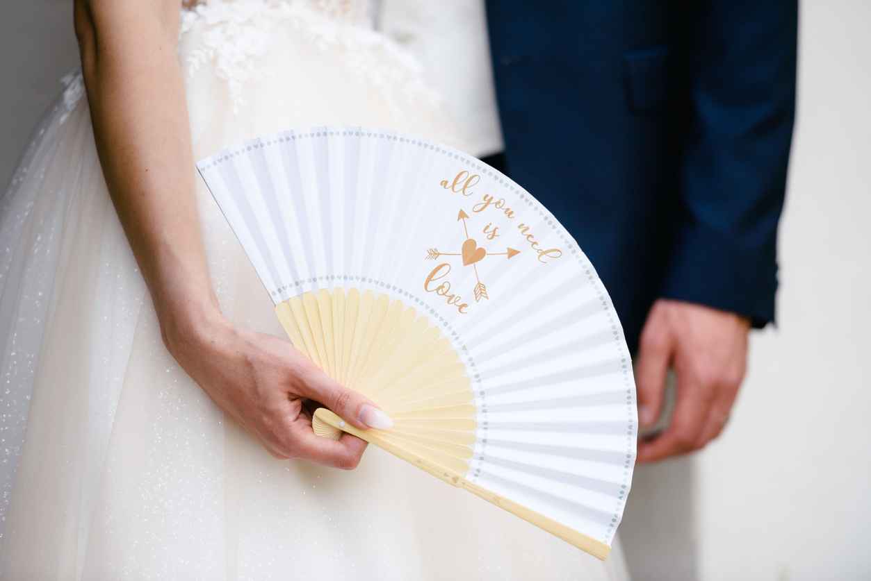 A closeup of a bride's hand holding a white fan standing besides bridegroom